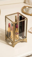 INVOKE decorative glass makeup brush holder with antique brass frame, ornate feet, holding lipsticks and mascaras.
