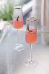 INVOKE fluted champagne flutes with gold rim and vertical ribs, filled with pink champagne and garnished with rosemary.