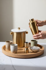 INVOKE luxury cocktail barware set with brushed gold shaker, ice bucket with marble lid, jigger, and cups on a wooden tray.