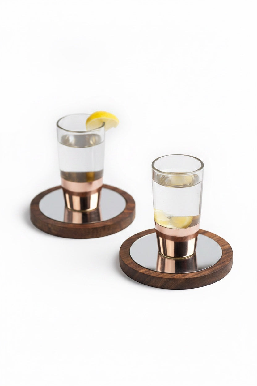INVOKE clear glass shot glasses with copper bands on wooden coasters with mirrored inserts, garnished with lemon slices.