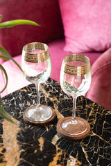 INVOKE elegant clear glass wine glasses with gold rim detail and wooden coasters on a marble table.